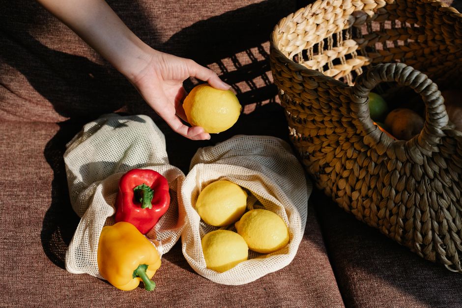 Crop anonymous person with lemon in hand standing near couch with ECO friendly bags with colorful bell peppers near wicker basket